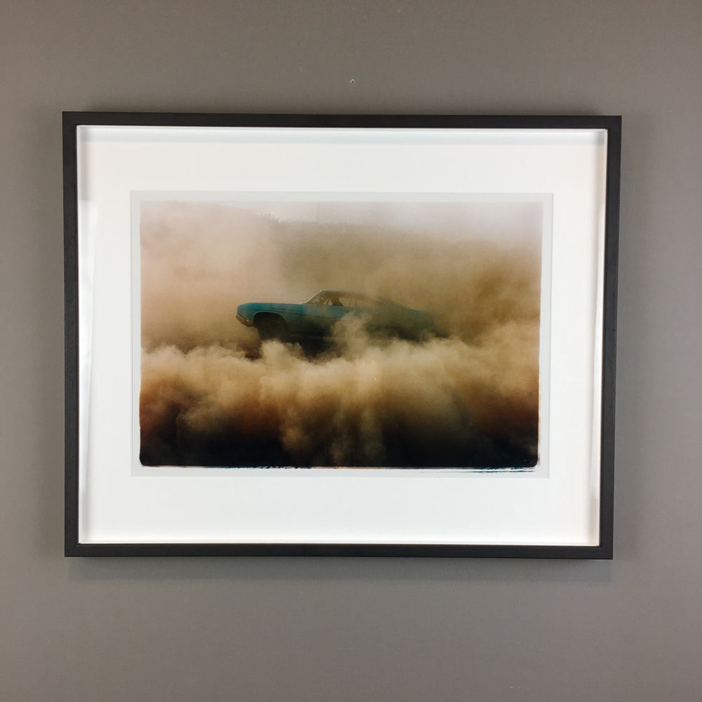 An image of a blue car driving through a dusty desert landscape on a white background, enclosed in a black frame